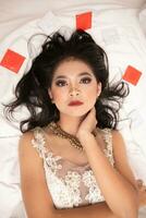un asiatique femme avec magnifique maquillage en train de dormir sur une blanc lit tandis que portant une robe photo