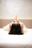 un asiatique femme dans une blanc robe est mensonge sur une blanc lit avec sa noir cheveux ample dans une Hôtel photo