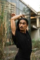 un asiatique homme avec longue cheveux et portant noir vêtements permanent autour un abandonné bâtiment photo