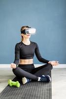 Jeune femme blonde en vêtements de sport portant des lunettes de réalité virtuelle méditant sur un tapis de fitness photo