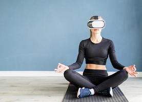 Jeune femme blonde en vêtements de sport portant des lunettes de réalité virtuelle méditant sur un tapis de fitness photo