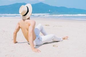 barebacked jeune homme asiatique assis sur le sable et regardant la mer photo