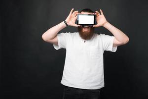 Portrait d'un homme barbu étonné tenant une tablette sur les yeux avec un écran blanc photo
