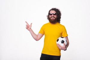 heureux bel homme barbu tenant un ballon de football et pointant vers l'extérieur photo