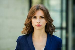 portrait de une magnifique Jeune femme avec longue marron cheveux, habillé dans une bleu veste, permanent contre le Contexte de un Bureau bâtiment. portrait de Jeune affaires femme en utilisant mobile téléphone photo