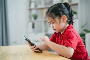 asiatique enfant fille à la recherche en utilisant et toucher mobile téléphone filtrer. bébé souriant marrant temps à utilisation mobile téléphone. aussi beaucoup écran temps. mignonne fille en train de regarder vidéos tandis que la télé dans vivant chambre, l'Internet dépendance. photo