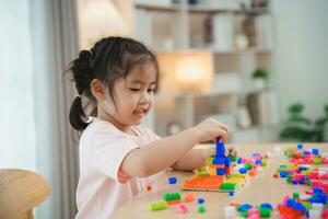 joyeux asiatique fille content et souriant en jouant coloré lego jouets, séance sur le table dans le vivant chambre, créative en jouant avec légo, bâtiment coloré les structures la créativité imaginer. photo