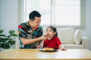 asiatique content les enfants fille et sa père alimentation en mangeant Pizza et souriant dans le vivant pièce à maison. les enfants fille et sa papa en mangeant et dégustation italien fait maison Pizza dans maison. photo