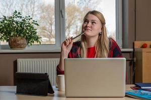 femme blonde souriante assise devant un ordinateur portable, buvant du thé et travaillant.