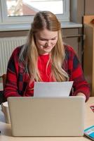 femme blonde souriante assise devant un ordinateur portable, buvant du thé et travaillant.