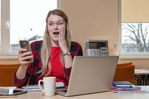 femme blonde souriante, assise devant un ordinateur portable et surprise, regarde le téléphone. photo