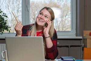 une femme blonde souriante assise devant un ordinateur portable et parlant au-dessus d'un téléphone portable. photo