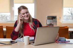 une femme blonde souriante assise devant un ordinateur portable et parlant au-dessus d'un téléphone portable. photo