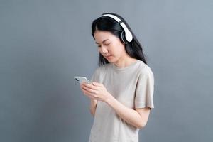 une femme asiatique porte un casque sans fil tenant un smartphone en regardant l'écran du téléphone à l'aide d'une application de lecteur mobile écoutant de la musique en ligne, apprenant une langue étrangère, regardant une vidéo se relaxant sur fond gris