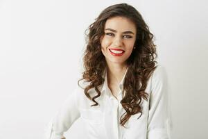 fermer portrait de souriant attrayant femme avec blanc dents, longue frisé cheveux, rouge rouge à lèvres maquillage à la recherche dans caméra isolé portant blanc chemisier photo