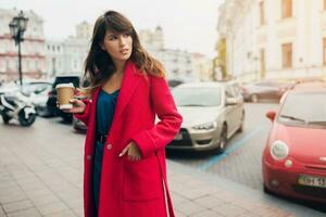 élégant femme dans rouge manteau en marchant dans rue avec café photo