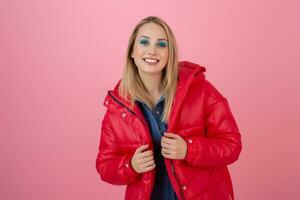 blond attrayant actif femme posant sur rose Contexte dans coloré hiver vers le bas veste de rouge couleur, ayant amusant, chaud manteau mode s'orienter, souriant photo