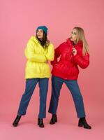 deux excité attrayant fille copains actif femmes posant sur rose Contexte dans coloré hiver vers le bas veste de brillant rouge et Jaune Couleur ayant amusement ensemble, chaud manteau mode tendance photo