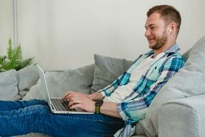 Beau souriant homme dans chemise séance détendu sur canapé à Accueil à table travail en ligne sur portable photo