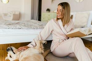 jolie souriant femme à Accueil dans Matin dans pyjamas en train de lire livre en jouant avec chien photo