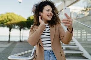 jolie frisé souriant femme en marchant dans ville rue dans élégant veste, en utilisant téléphone intelligent photo