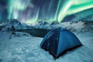 Tente bleue camping sur une colline enneigée avec des aurores boréales dansant sur une chaîne de montagnes sur l'île de Senja