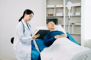 asiatique médecin dans blanc costume prendre Remarques tandis que discuter et asiatique âgé, homme patient qui mensonge sur lit avec recevoir saline Solution dans hôpital photo