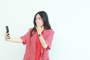 portrait de magnifique asiatique femme portant rouge tenue à la recherche à mobile téléphone avec surpris expression photo