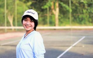 belle femme asiatique aux cheveux courts, portant un chapeau et souriant largement sur un court de tennis