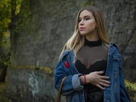 belle fille blonde modèle dans une veste en jean se tient dans la rue contre un mur de pierre dans le parc photo