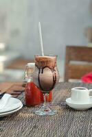 une à manger table avec une du froid Chocolat boisson servi dans une verre avec une inoxydable acier paille, le fusion Chocolat visible sur le verre ajoute à le délicieux effet. avec 2 bouteilles de ketchup photo