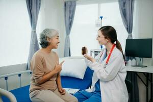 médecin dans blanc costume prendre Remarques tandis que discuter et asiatique âgé, femme patient qui mensonge sur lit avec recevoir saline Solution dans hôpital ou clinique. photo