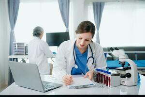 sur de soi Jeune caucasien femelle médecin dans blanc médical uniforme asseoir à bureau travail sur ordinateur. souriant utilisation portable écrire dans médical journal photo