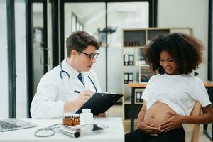 Enceinte africain femme a rendez-vous avec médecin à clinique. Masculin gynécologue ob gynécologue médical spécialiste avec stéthoscope écoute à de bébé battement de coeur dans de la mère ventre. photo