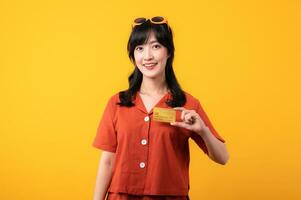 portrait Jeune magnifique asiatique femme content sourire habillé dans Orange vêtements montrant crédit carte isolé sur Jaune Contexte. Payer et achat achats Paiement concept. photo