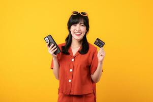 portrait Jeune magnifique asiatique femme content sourire habillé dans Orange vêtements en utilisant crédit carte achat achats en ligne sur téléphone intelligent isolé sur Jaune Contexte. Payer et achat Paiement concept. photo