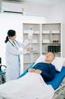 asiatique médecin dans blanc costume prendre Remarques tandis que discuter et asiatique âgé, homme patient qui mensonge sur lit avec recevoir saline Solution dans hôpital photo