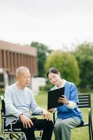 personnes âgées asiatique Sénior homme sur fauteuil roulant avec asiatique prudent soignant et encourager patient, en marchant dans jardin. avec se soucier de une soignant et Sénior santé assurance. photo