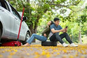 asiatique couple en utilisant téléphone intelligent pour assistance après une voiture panne sur rue. concept de véhicule moteur problème ou accident et urgence Aidez-moi de professionnel mécanicien photo