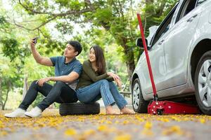 asiatique couple en utilisant téléphone intelligent pour assistance après une voiture panne sur rue. concept de véhicule moteur problème ou accident et urgence Aidez-moi de professionnel mécanicien photo