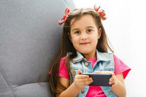 mignonne peu fille en utilisant une téléphone intelligent, à la recherche à caméra et souriant tandis que sur le canapé à Accueil photo