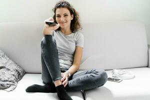 portrait de souriant Jeune femme séance sur le canapé en utilisant éloigné contrôle photo