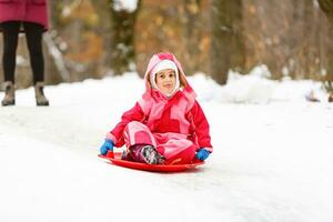 mignonne peu fille glissement vers le bas de le colline Haut couvert avec blanc et duveteux neige. un de le les enfants préféré activité dans hiver temps. photo