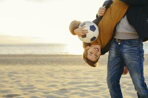 père en portant fils avec Football à l'envers vers le bas sur le plage à le coucher du soleil photo