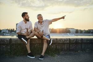 père et adulte fils séance sur une mur à le au bord de la rivière à le coucher du soleil en buvant une Bière photo