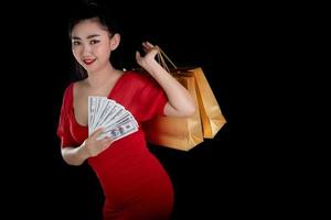 portrait d'une belle jeune femme asiatique vêtue d'une robe rouge tenant un billet de 100 USD et des sacs à provisions sur fond noir, regardez la caméra photo