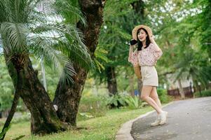 portrait de asiatique Jeune femme voyageur avec tissage chapeau et panier content sourire sur vert Publique parc la nature Contexte. périple voyage mode de vie, monde Voyage explorateur ou Asie été tourisme concept. photo