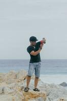 indonésien Masculin photographe portant une chapeau et noir T-shirt permanent sur corail rochers avec une plage vue dans le Contexte. Extérieur actif vacances aventure Voyage concept. photo