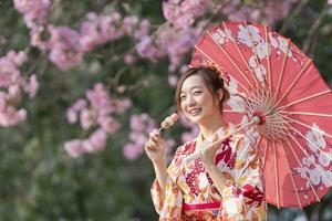 Japonais femme dans traditionnel kimono robe en portant parapluie et sucré hanami dango dessert tandis que en marchant dans le parc à Cerise fleur arbre pendant le printemps Sakura Festival avec copie espace photo