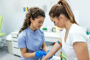 préparation pour du sang tester avec jolie Jeune femme par femelle médecin médical uniforme sur le table dans blanc brillant chambre. infirmière perce le les patients bras veine avec aiguille Vide tube. photo